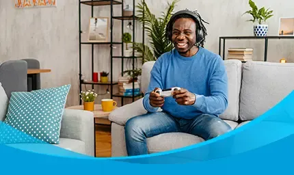 A smiling man wearing headphones plays a video game with a controller on his couch.